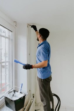 Mavi tişörtlü, koyu renk sakallı, genç beyaz bir adam pencerenin kenarında duruyor ve tavanın altındaki duvarın köşelerini beyaz boya ile boyuyor..