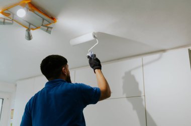 Koyu renk sakallı, mavi tişörtlü genç bir beyaz adam arkasında duruyor ve tavanda bir silindir ile beyaz boya çiziyor. Avizenin yanında mutfak dolapları asılı, alt tarafı seçici odaklı..