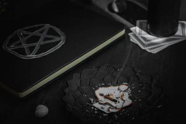 stock image A black metal saucer with creased paper with a wisp of smoke, a leather book with a magical white symbol, black candles and stones lie on a black wooden background, close-up side view. Esoteric