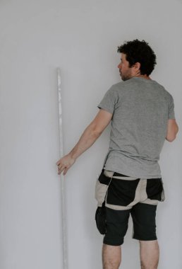 A young handsome caucasian plasterer in a gray t-shirt, work pants and with brown curly hair holds a large spatula with his hand and checks the evenness of the wall after puttying, close-up side view