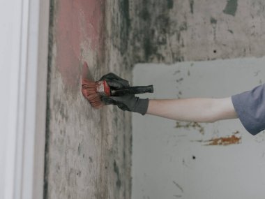 Eldivenli tanınmayan genç bir adam kirli bir duvara fırçayla kırmızı çimento sürüyor. Küflü lekeler, yakın çekim. Ev tadilatı ve inşaat işi kavramı.