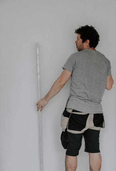 A young handsome caucasian plasterer in a gray t-shirt, work pants and with brown curly hair holds a large spatula with his hand and checks the evenness of the wall after puttying, close-up side view