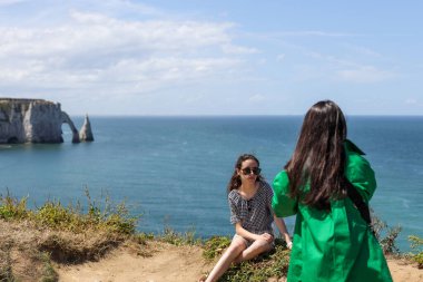 Esmer, dalgalı, uzun saçlı bir kız yeşil tişörtlü, arkasında duruyor ve genç kız kardeşinin deniz manzaralı bir dağın tepesinde poz verirken ve otururken çekilmiş bir fotoğrafını çekiyor.