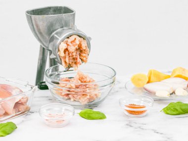 Elle öğütülmüş et öğütücüsü, cam kasede kıyma, kemiksiz tavuk butları, pembe tuz, soğan kabuğu, çay tabağında sarımsak ve mermer bir masada beyaz bir duvara karşı fesleğen yaprakları.