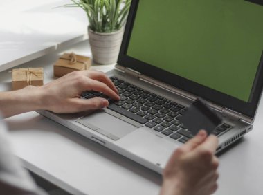 Gri tişörtlü genç beyaz bir adam online alışveriş yapıyor çalışma masasında bir koltukta dizüstü bilgisayarla oturuyor ve elinde bir kredi kartı tutuyor. Çevrimiçi alışveriş konsepti.
