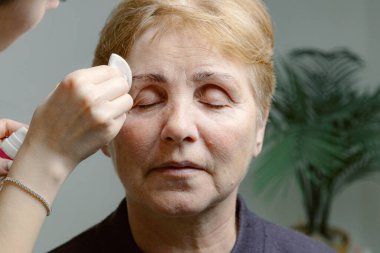 Genç, beyaz, tanınmayan bir kozmetik uzmanı yaşlı bir kadının sağ kaşındaki boyayı pamuk peçeyle temizleyici kullanarak dikkatlice çıkarıyor.