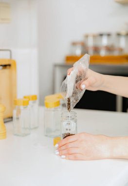 One young Caucasian unrecognizable girl pours seasoning spice black peppercorns from a plastic bag into a new glass transparent jar, standing at a white table in the kitchen on a summer day, side view