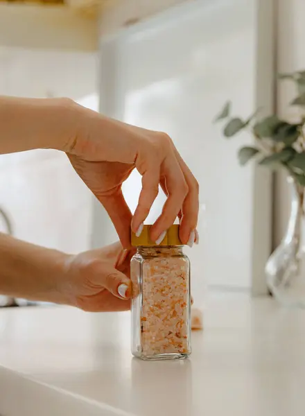 stock image One young Caucasian unrecognizable girl closes a glass jar with the seasoning spice pink salt with a wooden lid, standing at a white table in the kitchen on a summer day, side view close-up with depth