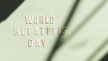 Dünya Hepatit Günü cümlesine sahip hafif ahşap harfler solda, atmosferik soyut gölgeler ve sağda düz bir kopya alanı olan nane yeşili bir zemin üzerinde uzanıyor. Dünya