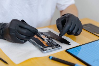 Beyaz tişörtlü ve siyah lateks eldivenli tanınmayan genç bir beyaz adam, cımbız kullanarak bir sürgüyü akıllı telefona yerleştirir, bir yaz günü mutfaktaki bir masada oturur, yakın çekimde yan görüş sağlar.