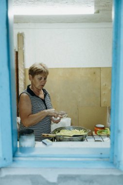 Yaşlı, beyaz, güzel bir kadın yaz günü eski bir kır evinin sobasında tavada kızaran ellerinde patateslerle tuzla birlikte mavi çerçeveli bir pencereden içeri bakıyor.