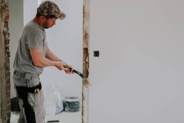 Üniformalı ve gri tekstil eldivenli genç beyaz bir adam levyeyle yarım yamalak duran levhayı söküyor kapıdaki eski alçıyı söküyor ve sağında fotokopi alanı var.