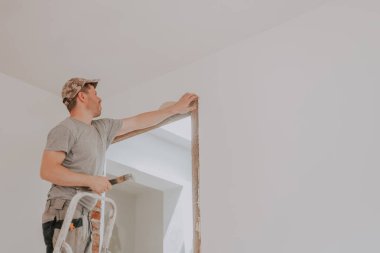 Üniformalı ve şapkalı genç bir beyaz adam, beyaz duvarlı bir odada merdiven üzerinde dururken, aşağıdan seçici bir odak noktasıyla, eski macunu kullanarak kapı aralığını temizliyor.