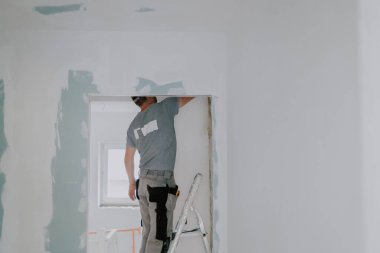 Gri tişörtlü genç bir beyaz adam, bir eliyle baltayla kapı aralığındaki eski macunu temizliyor. Bir yandan merdivenin kenarında dikilirken, diğer yandan seçici bir odak noktasıyla.