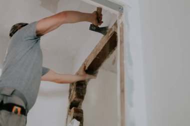 Gri tişörtlü ve şapkalı beyaz bir genç kapı aralığındaki eski macunları temizlemek için balta kullanıyor. Enkaz dökülüyor, merdivenin üstünde duruyor, aşağıdan seçici bir bakış açısı var.