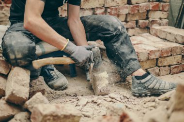 A young caucasian man in black work clothes sits on a small chair on the left and cleans the bricks from dirt, debris and old cement with an ax, side view close-up.Construction work concept.