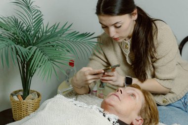 Beyaz tenli, genç, güzel, esmer bir kozmetik uzmanı, yatmakta olan yaşlı bir müvekkil için katman işlemine başlamadan önce akıllı bir telefonla sağ gözünün kirpiklerinin fotoğrafını çekiyor.