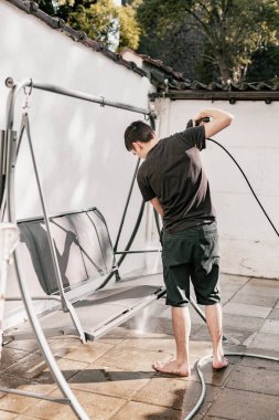 Siyah elbiseli, esmer, beyaz bir adamın portresi. Çıplak ayakla evinin arka bahçesinde salıncağını yıkıyor..