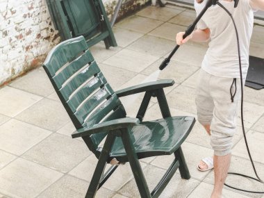 Gri eşofmanlı ve tişörtlü beyaz bir adam evinin arka bahçesindeki koyu yeşil güneş musluğunun altında plastiği yıkıyor. Güneş ışığı, ev yıkama konsepti.