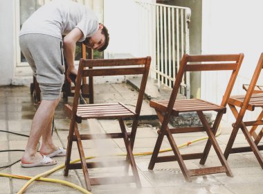 Gri eşofmanlı ve tişörtlü beyaz bir adam evinin arka bahçesindeki Karcher ahşap bahçe sandalyelerinden gelen donmuş spreyle yıkanıyor. Bahçeyi temizleme konsepti