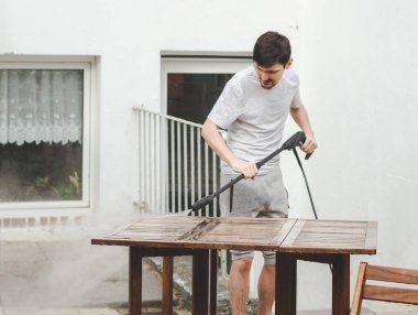 A caucasian guy in gray sweatpants washes a wooden garden table in the backyard of his house with frozen spray from a karcher, close-up. The concept of cleaning the yard, preparing for the holiday, at clipart