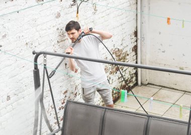 Gri eşofmanlı ve tişörtlü yakışıklı beyaz bir adam evinin arka bahçesindeki, yan manzaralı, bir Karcher 'ın baskısı altında üçlü hasır bir bahçe salıncağını yıkıyor. Konsept