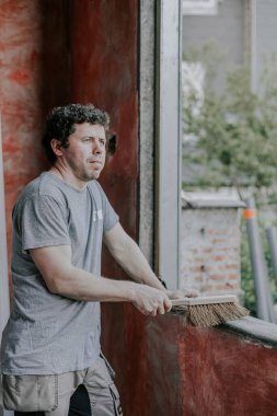 Yaşlı bir süpürge tutan genç beyaz bir adam düşünceli bir şekilde yan tarafa bakıyor kirli bir pencere çerçevesini süpürüyor, yakından yan görüş. Pencereleri temizleme ve kurma konsepti, inşaat işi, ev