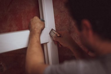 Kıvırcık saçlı genç beyaz bir adam pencere kenarındaki çatlakları inşaat bıçağıyla temizliyor ve onları montaj için hazırlıyor. Ev yenileme, montaj konsepti