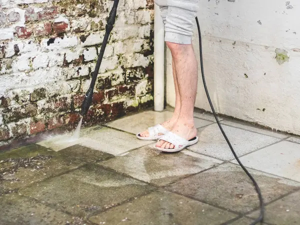 stock image A caucasian man with hairy legs in white slippers washes the stone tiles of the terrace near the old painted brick wall in the backyard of his house under the pressure of water from the kercher, close