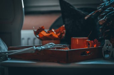 Kahveli ahşap tepsi, tabakta waffle, sepette kavak yaprakları, hayalet gözlükleri, yanan bir çelenk, cadı şapkası ve komodine uzanan bir iskelet el, yakın çekim görüntüsü.