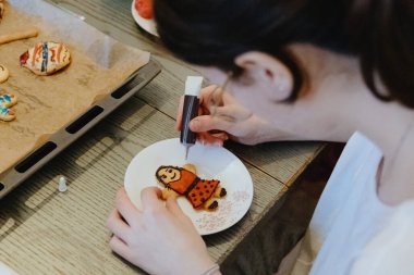 Beyaz tişörtlü güzel bir beyaz kız bir masada oturuyor, pişmiş kurabiyeleri bir tüp çikolatayla dekore ediyor, koyu gri bir masada oturuyor, yakın plan yan görüş. Adım adım talimatlar
