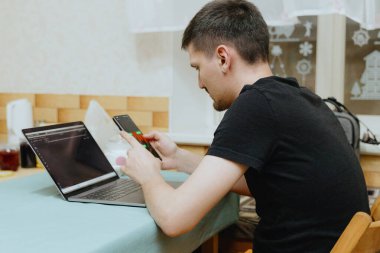 Beyaz tenli yakışıklı bir adamın portresi mutfaktaki masada yan otururken aynı anda hem laptop hem de akıllı telefon üzerinde çalışıyor.