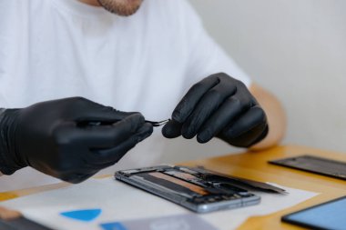 Beyaz tişörtlü ve siyah lateks eldivenli yakışıklı beyaz bir adam parmaklarıyla akıllı telefonu tamir etmek için tuttuğu cıvatayı alır ve bir yaz günü mutfakta bir masada oturur.