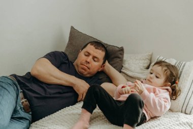 Genç, beyaz, yakışıklı bir adamın portresi. Baba ve küçük bir kız. Kanepede birlikte yatıyorlar ve oturma odasında hevesle televizyon izliyorlar.