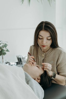 Ofisteki kozmetoloji masasında uzanan kadın bir müşterinin solunda iki cımbızla yapay kirpiklerini uzatan güzel beyaz tenli bir kozmetik uzmanının portresi.