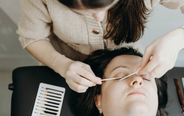 Genç ve güzel bir beyaz kadın kozmetik uzmanının portresi. İki çubukla dikkatlice temizler. Kozmetoloji masasında uzanan bir bayan müşterinin sağ gözündeki doğal kirpikleri.