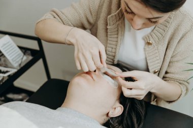 Beyaz tenli güzel kozmetik uzmanı bir kızın portresi. Ofisteki kozmetoloji masasında uzanan bir kadın müşterinin sol göz kapağının altına kağıt yapıştırıcı yapıştırıyor.