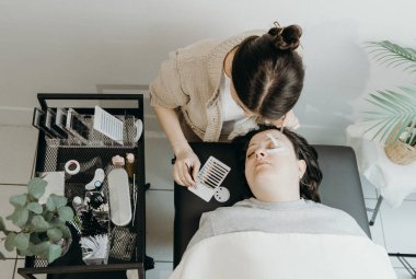 Beyaz tenli güzel bir kızın portresi, kozmetik uzmanı yapay kirpiklerini kadın bir müşterinin sol gözünde kozmetoloji masasında uzanan cımbızla yapıştırıyor.