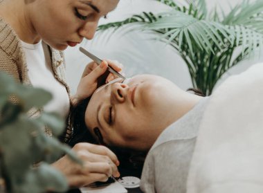 İki cımbız kullanarak dikkatle yapay kirpiklerini yapıştıran güzel beyaz bir kız kozmetik uzmanının portresi. Evdeki ofisinde kozmetoloji masasında yatan kadın bir müşteri.