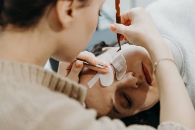 Beyaz tenli güzel bir kozmetik uzmanı kızın portresi. Kozmetolojide yatan bir kadın müşterinin sol gözkapağının alt kısmındaki iki cımbızla kağıt etiketlerini soyuyor.