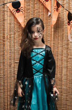 Yüzünde Cadılar Bayramı makyajı olan beyaz bir kızın portresi hasır bir çitin arkasında duruyor. Bayram bayraklarıyla kameraya bakıyor.