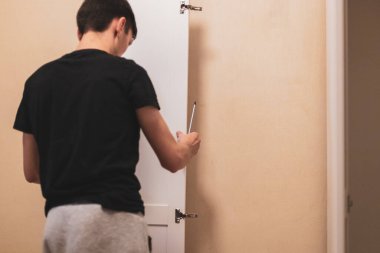 Arkadan duran beyaz tenli genç adam, montaj için duvarın yanındaki dolabın yan kapısını kullanıyor, seçici bir odak ile yakın plan yan görüş. Mobilya montaj konsepti.