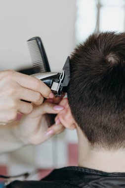 Tanınmayan genç bir kuaförün elleri sol şakak bölgesinde saç kesiyor, kulağını bir parmak ile tutuyor ve yaz günü evde esmer bir adama saç makasıyla vuruyor.