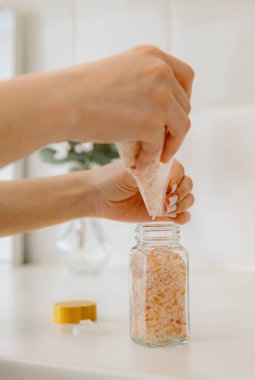 Tanımlanamayan genç bir beyaz kız plastik bir torbadan yeni bir cam şeffaf kavanoza pembe tuz çeşnisi döker, yaz günü mutfakta beyaz bir masada durur, yan görüş yakındır.