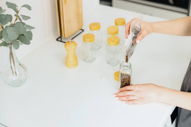 Tanınmayan genç bir beyaz kız plastik bir torbadan baharatlı siyah biberleri yeni bir cam şeffaf kavanoza döküyor. Yazın üst tarafında, mutfakta beyaz bir masada duruyor.