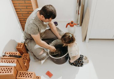 Üniformalı genç beyaz bir adam, küçük kızıyla birlikte, spatulayla bir kovada çimento harcı yoğuruyor, şömine için sigara içen bir adamın onarılıp yerleştirildiği bir odada çömeliyor.