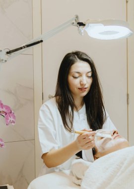 Genç, beyaz tenli, güzel bir kozmetik uzmanı gülümseyerek aşağıya bakar ve beyaz krem bir maske takar. Burun ve dudakların olduğu bölgede, yetişkin bir müşterinin yüzüne fırça atar.