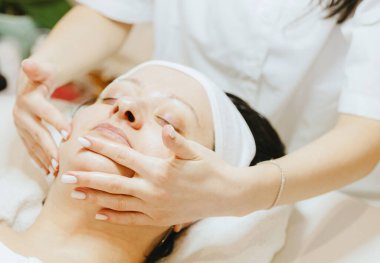 Tanımlanamayan genç bir kozmetik uzmanı, gözleri masaj masasına kapalı bir şekilde uzanan yetişkin bir müşteriye parmaklarıyla nazikçe çene ve yanaklarına masaj yapar.