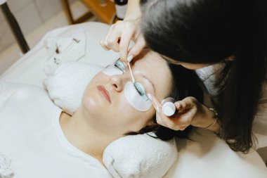 Beyaz önlüklü, yarı tanınmış beyaz bir kozmetik uzmanı kızın portresi. Sol gözünün kirpiklerindeki kahverengi boyayı çıkartıyor. Pamuklu çubukla esmer bir kadına uzanıyor.