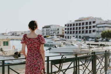 A portrait of one young beautiful Caucasian brunette woman in a bright colorful dress stands with her back to the left, leaning against an iron railing against the backdrop of a blurred embankment